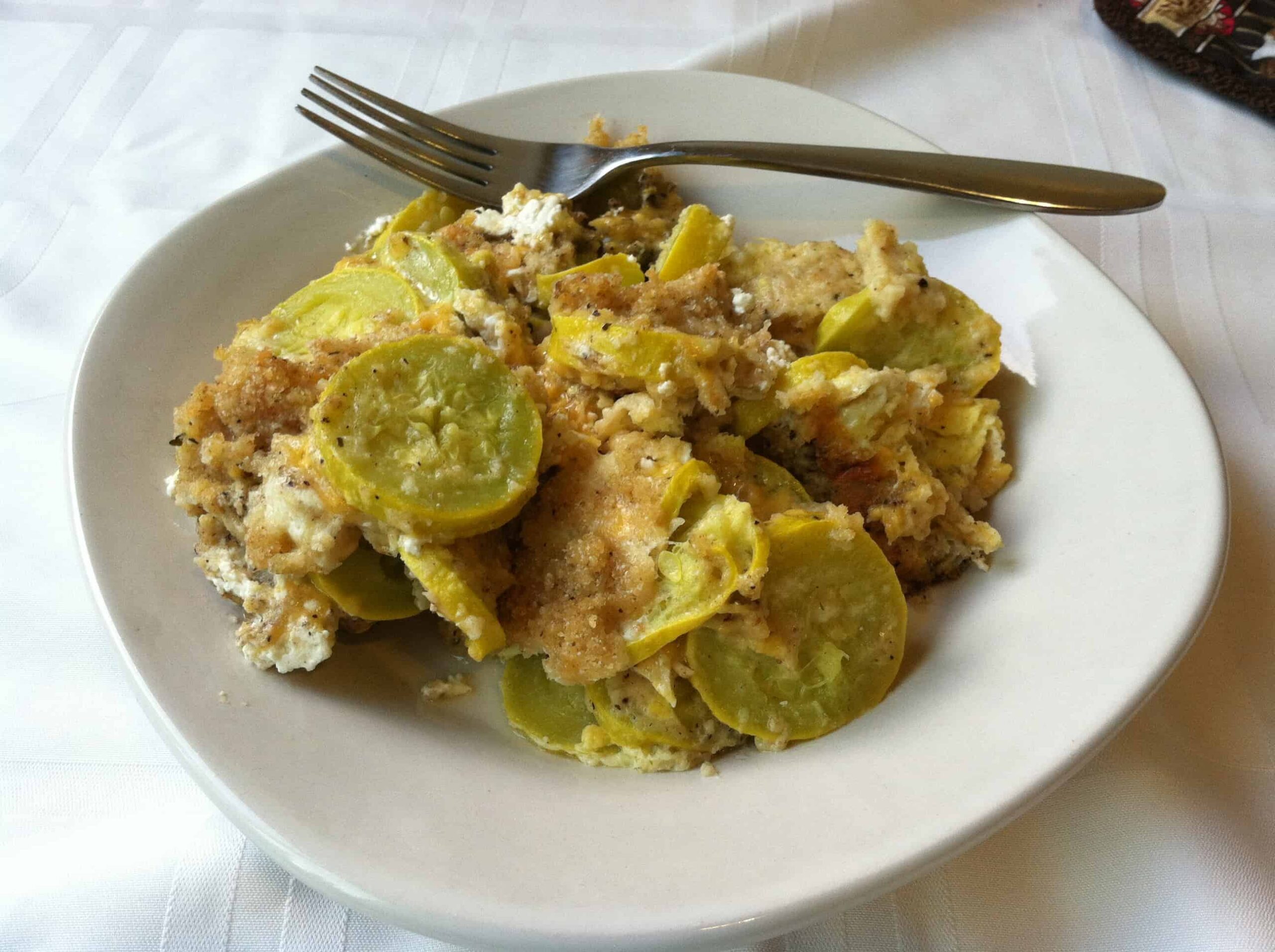 A white plate holds a serving of two cheese squash casserole, topped with breadcrumbs and cheese. A fork rests on the plate. The casserole appears baked and golden brown, with a mix of yellow squash and creamy filling. The background features a white tablecloth, completing this delightful recipe scene.