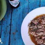 A hearty bowl of chicken and sausage gumbo with shredded meat, vibrant vegetables, and flavorful sausage sits on a blue wooden table. Nearby, a bottle of sauce and a green plate add to the scene. A wooden spoon and a partial 3 Boys and a Dog logo complete this inviting setting.