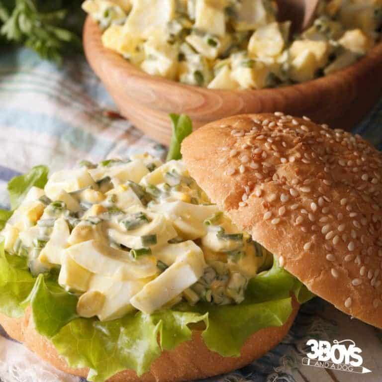 A sandwich with curried egg salad and lettuce on a sesame seed bun takes center stage, while a wooden bowl brimming with more curried egg salad complements the scene. The sandwich rests on a patterned cloth. In the corner, a logo reads 3 Boys & a Dog.