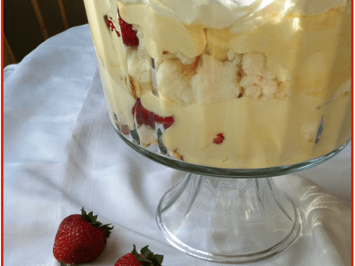 A layered dessert in a glass bowl showcases whipped cream, sliced strawberries, and cake pieces. The bowl rests on a white cloth with whole strawberries scattered around. Text at the top reads Easy Strawberry Trifle Recipe, and 3 Boys and a Dog graces the bottom.