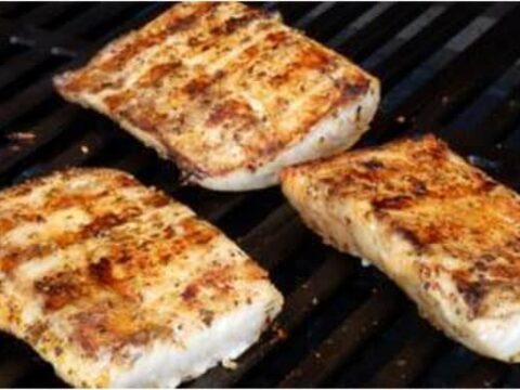Three pieces of seasoned Simple Ginger Lime Mahi Mahi are being grilled on a barbecue grill, showing a golden-brown sear with grill marks. The fillets are positioned closely on the black metal grates.