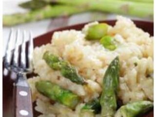 A plate of Risotto with Asparagus, featuring creamy goodness mingling with tender asparagus pieces, is accompanied by a fork. In the background, fresh asparagus stalks are elegantly displayed, echoing the dishs vibrant flavors.