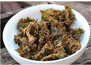 A white bowl filled with Spicy “Cheesy” Kale Chips sits on a wooden surface. The crispy chips boast a tantalizing green and brown appearance, perfectly hinting at their bold seasoning blend that delivers a zesty kick and irresistible cheesy flair.