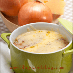 A green bowl of French Onion Soup, adorned with melted cheese, rests on a white saucer with a spoon. Background details include whole onions and a wedge of cheese. French Onion Soup is prominently displayed at the top, accompanied by a website URL below.