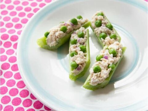 Plate with celery stalks filled with a tuna canoe salad mixture, garnished with green peas and red onions. The plate is on a pink and white polka dot tablecloth. FamilyFun magazine is printed in the top left corner.