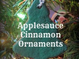 A homemade green tree-shaped ornament dangles on a Christmas tree adorned with colorful lights, spreading holiday cheer. The text reads Applesauce Cinnamon Ornaments, inviting you to add a sweet fragrance to your festive decor.