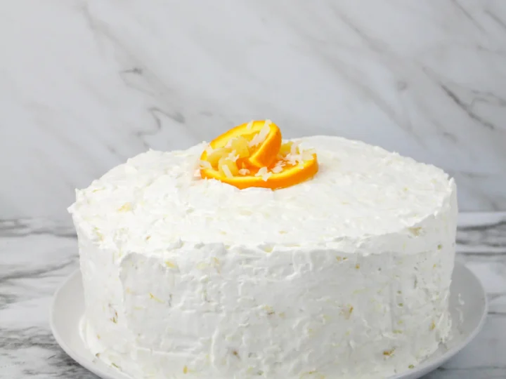 A round cake with white frosting and a touch of coconut flair, topped with orange slices, sits on a white plate. The background features a marble surface.