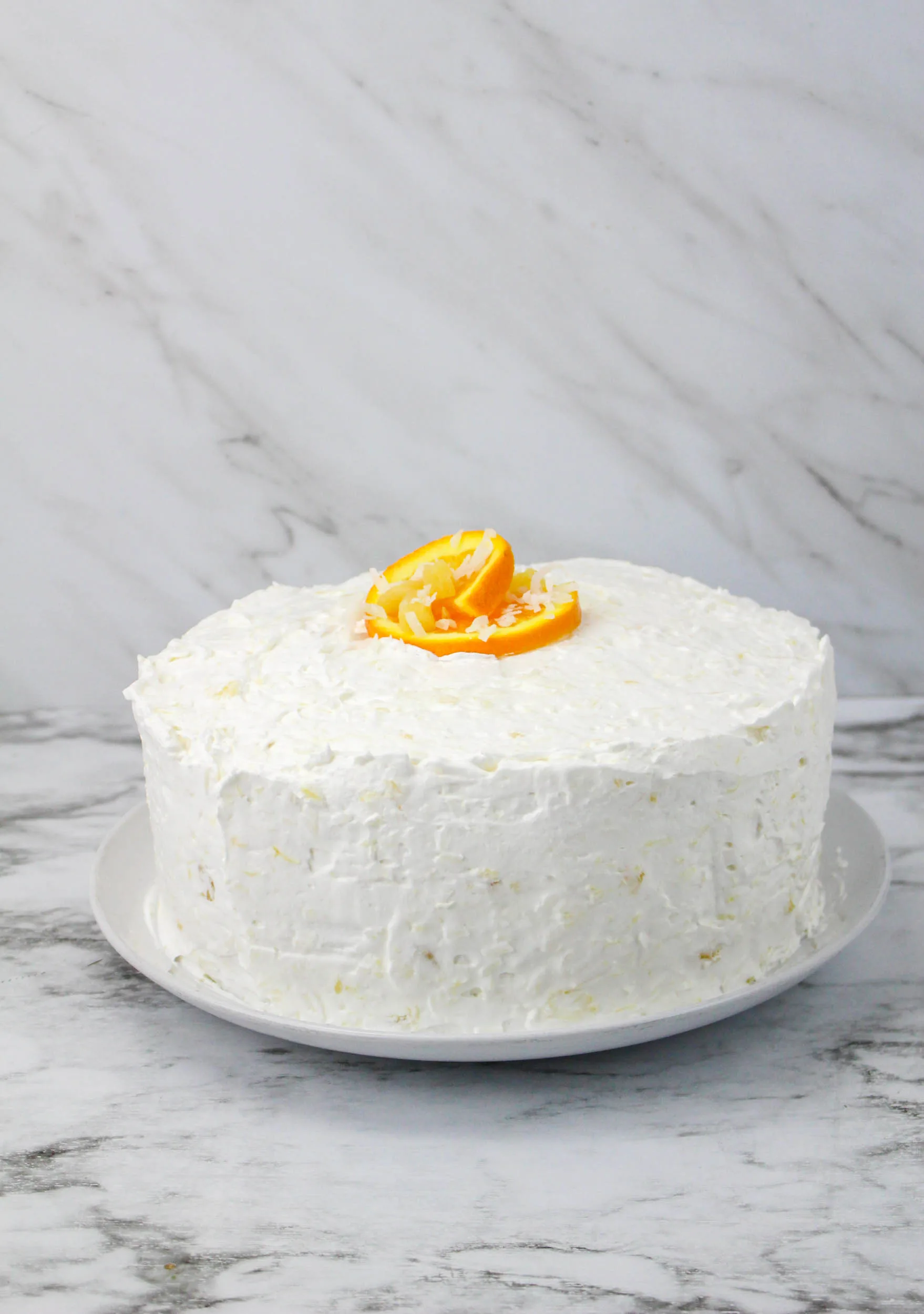 A round cake with white frosting and a touch of coconut flair, topped with orange slices, sits on a white plate. The background features a marble surface.