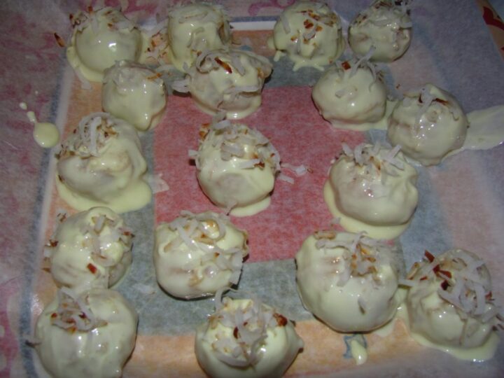 Fourteen white chocolate-covered truffles, perfect for coconut recipes, topped with shredded coconut on parchment paper. Some truffles feature slightly melted chocolate, and the coconut is sprinkled unevenly.