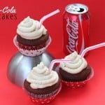 Three chocolate cupcakes with light brown frosting and red straws sit elegantly on a metallic stand against a vibrant red background. Beside them, a can of Coca-Cola completes the scene. The words Coca-Cola Cupcake Recipe grace the top left corner, inviting you to indulge.