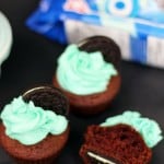 Three chocolate cupcakes with bright blue frosting, each topped with a Chocolate Mint Oreo, tempt the senses. One cupcake is bitten, revealing the rich interior. A package of cookies is partially visible in the background, hinting at a delightful recipe to try at home.