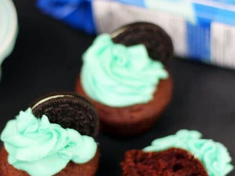 Three chocolate cupcakes with bright blue frosting, each topped with a Chocolate Mint Oreo, tempt the senses. One cupcake is bitten, revealing the rich interior. A package of cookies is partially visible in the background, hinting at a delightful recipe to try at home.