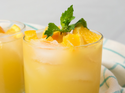 A refreshing mocktail in a short glass filled with orange juice, ice cubes, and garnished with orange slices and a sprig of mint. Perfect for kids parties, the drink sits on a light-colored surface next to a white and green checkered cloth.