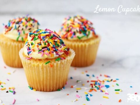 Three lemon cupcakes with frosting and colorful sprinkles are arranged on a marble surface, perfect for any Lemon Cupcake Recipe. More sprinkles are scattered around. The text Lemon Cupcakes graces the top right corner, adding a sweet touch.
