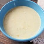 A blue bowl filled with creamy yellow soup sits invitingly on a wooden surface, hinting at the comfort of an easy broccoli braid recipe waiting nearby. A beige cloth napkin is partially visible next to the bowl, completing this cozy scene.