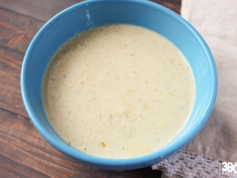 A blue bowl filled with creamy yellow soup sits invitingly on a wooden surface, hinting at the comfort of an easy broccoli braid recipe waiting nearby. A beige cloth napkin is partially visible next to the bowl, completing this cozy scene.