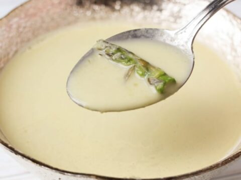 A silver spoon holds a creamy soup with asparagus tips over a textured silver bowl filled with the same soup. In the background, a light, blurred surface complements this cozy dish, much like enjoying an Easy Broccoli Braid Recipe on a lazy afternoon.
