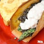 A close-up of mouthwatering lentil & asparagus tacos, each crispy shell generously stuffed with lentils and tender asparagus, topped with a dollop of creamy sauce, all set on a vibrant red plate.