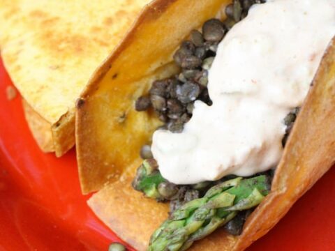 A close-up of mouthwatering lentil & asparagus tacos, each crispy shell generously stuffed with lentils and tender asparagus, topped with a dollop of creamy sauce, all set on a vibrant red plate.