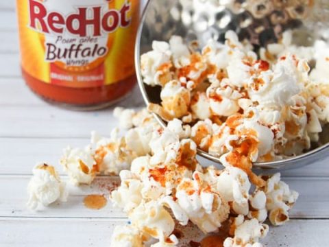 A bowl of Buffalo-style hot popcorn spills onto a white surface, drizzled with spicy goodness. In the background, a bottle of Franks RedHot Buffalo Wing Sauce stands proudly, ready to elevate your snacking game with every fiery bite.