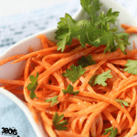 A close-up of a simple carrot salad in a white bowl, featuring julienned carrots garnished with fresh parsley. The dish rests on a light blue and white cloth.