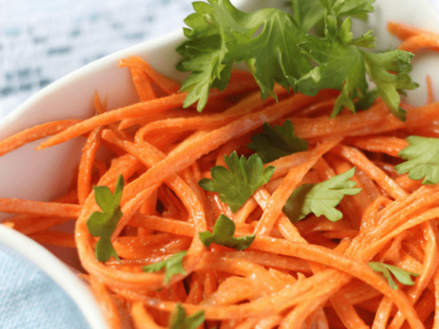 A close-up of a simple carrot salad in a white bowl, featuring julienned carrots garnished with fresh parsley. The dish rests on a light blue and white cloth.