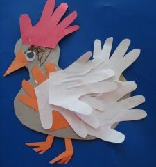 Set against a blue background, this Alabama crafts for kids project features a colorful chicken made from cut-out paper hand shapes. It boasts a pink hand-shaped comb, white and orange feathers, a brown body, an orange beak and feet, and a googly eye.
