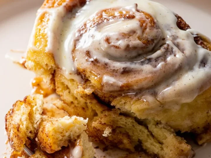 A close-up of a cinnamon roll, one of my favorite bread recipes, sits on a beige plate, topped with creamy icing. A fork rests nearby, holding a piece of the roll. The layers of dough and cinnamon filling are tempting and deliciously visible.
