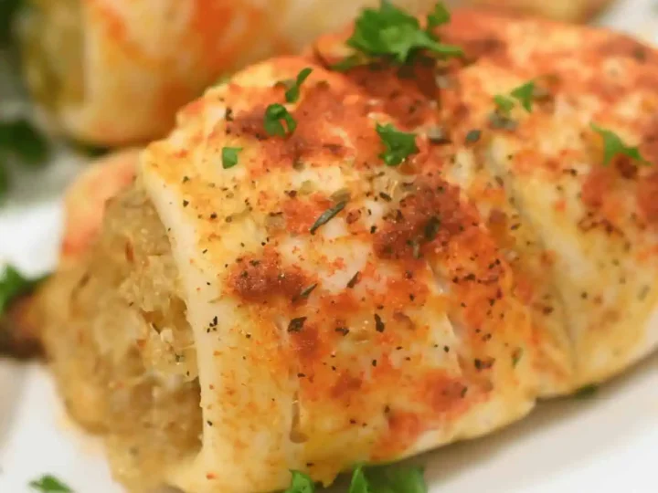 Close-up of a cooked stuffed chicken breast on a white plate. The chicken, reminiscent of healthy tilapia recipes, is seasoned with herbs and paprika, and garnished with fresh parsley. Another portion is visible in the background.