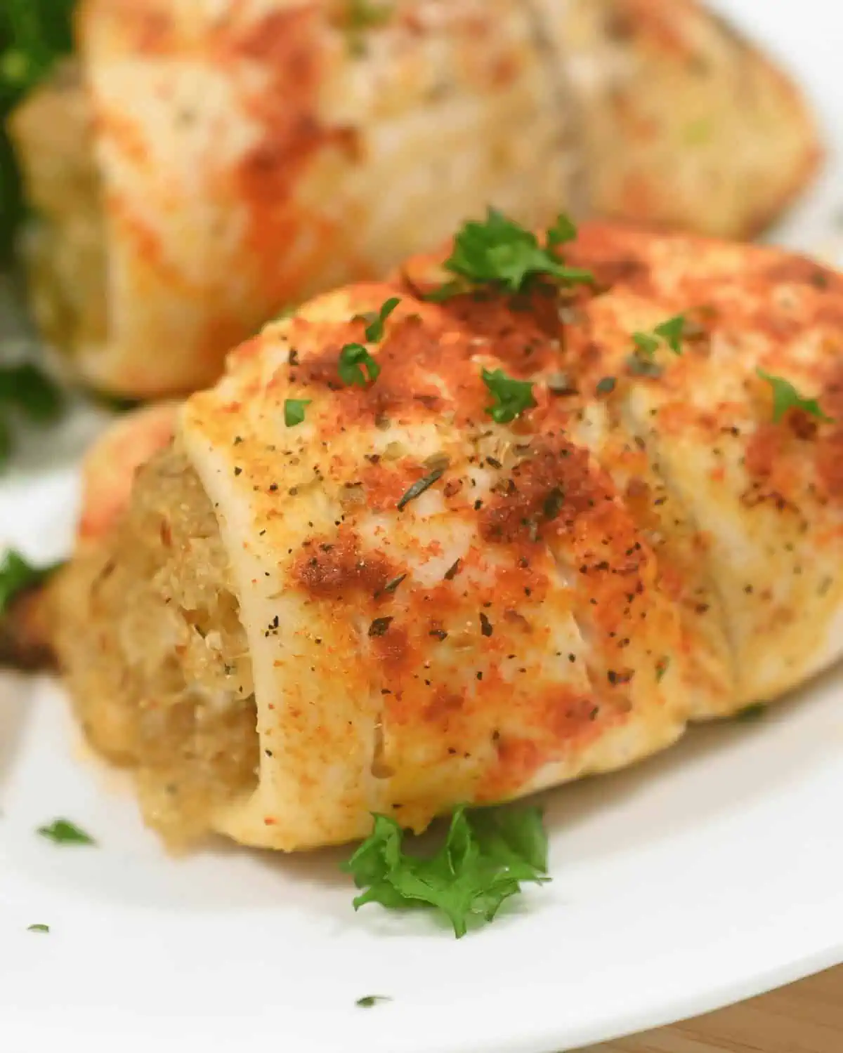 Close-up of a cooked stuffed chicken breast on a white plate. The chicken, reminiscent of healthy tilapia recipes, is seasoned with herbs and paprika, and garnished with fresh parsley. Another portion is visible in the background.