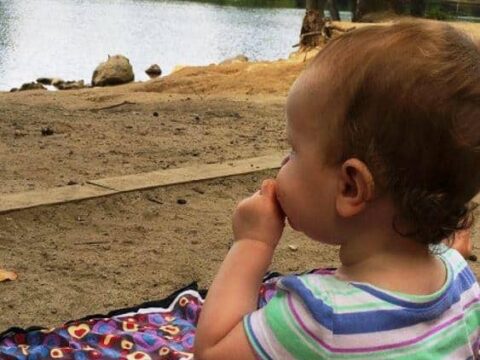 A toddler in a striped shirt and pink shorts sits on a blanket by a lake, enjoying a fruit snack. The tranquil scene, surrounded by trees and clear skies, highlights some of the best kid-friendly activities in Western Mass.
