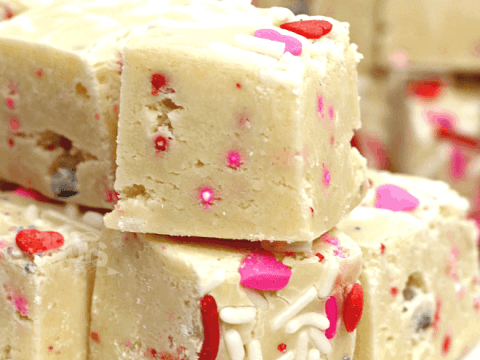 Image of several pieces of Valentine's Day cake mix fudge stacked on a plate. The fudge is decorated with pink, red, and white sprinkles, perfectly capturing the Valentine's spirit. The text reads Cake Batter Fudge Valentine's Day Recipe.
