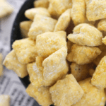 A close-up of a bowl filled with square, yellow, cheese-flavored snacks. The background reveals a blurred section of a newspaper. A few snacks, alongside some Pumpkin Spice Puppy Chow, are scattered around the bowl, adding an autumnal twist to the scene.