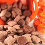 A pile of chocolate-covered snacks, including Reeses Peanut Butter Cup Puppy Chow, spills from an orange bowl onto a white surface. Small peanut butter cups are mixed in with the treats, and several wrapped chocolates are visible on the side.