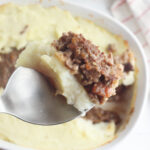 A close-up of a spoon holding a piece of shepherds pie, reminiscent of a hearty meatloaf casserole recipe. The dish is topped with creamy mashed potatoes and minced meat in a white baking dish, with a cloth featuring red stripes in the background.