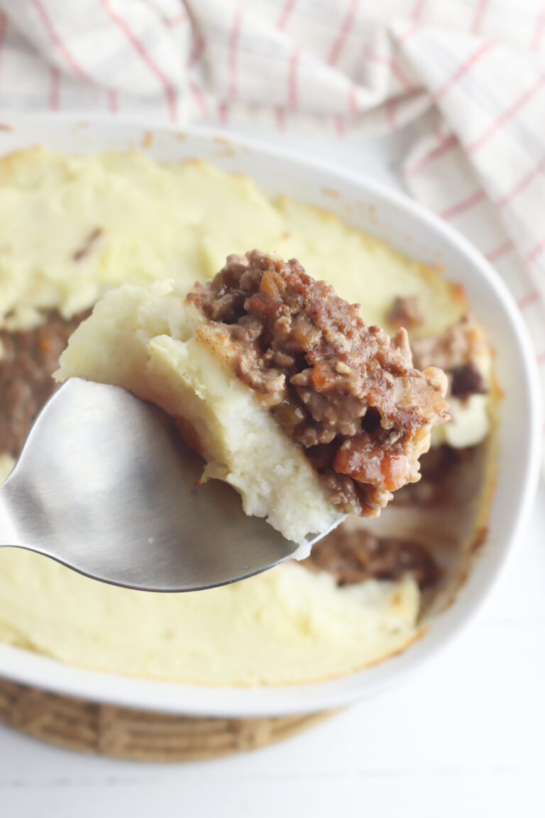 A close-up of a spoon holding a piece of shepherds pie, reminiscent of a hearty meatloaf casserole recipe. The dish is topped with creamy mashed potatoes and minced meat in a white baking dish, with a cloth featuring red stripes in the background.