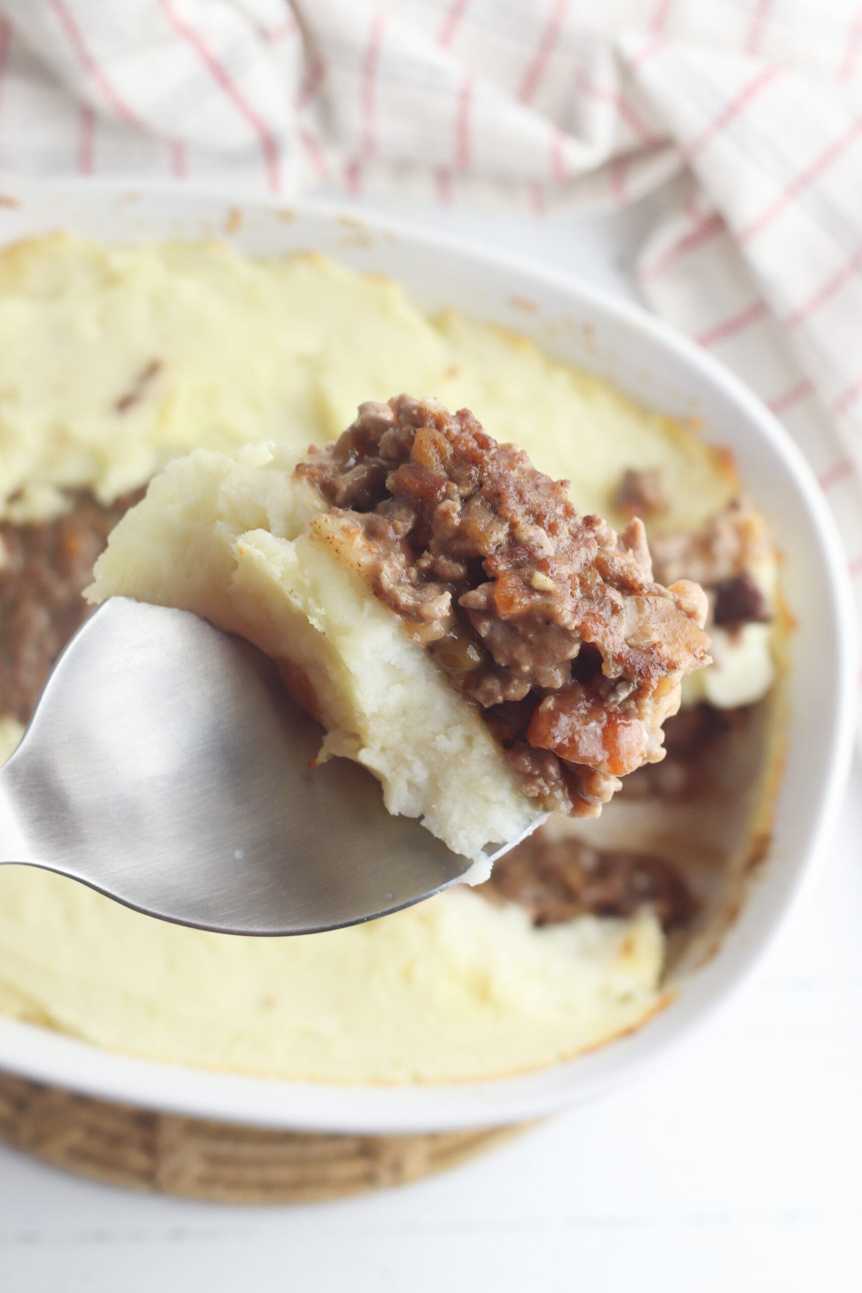 A close-up of a spoon holding a piece of shepherds pie, reminiscent of a hearty meatloaf casserole recipe. The dish is topped with creamy mashed potatoes and minced meat in a white baking dish, with a cloth featuring red stripes in the background.