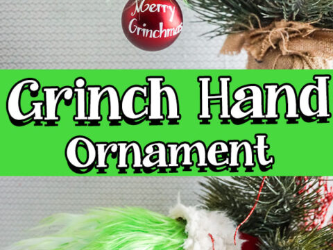 A festive ornament resembling the Grinchs green hand, perfect for those wondering how to make a Grinch hand, holds a red bauble with Merry Grinchmas on it. It hangs on a Christmas tree branch against a vibrant green background displaying Grinch Hand Ornament.