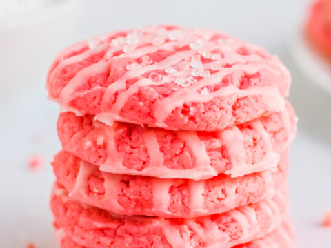 A stack of six pink cookies made from a strawberry cake mix sits enticingly on a white surface, drizzled with white icing and sprinkled with sugar crystals. The blurred background adds an air of mystery to this delectable treat.