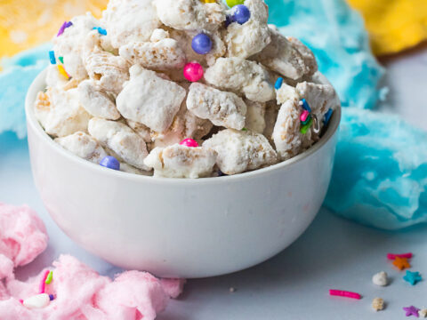 A delightful bowl of powdered sugar-covered snack mix, topped with colorful sprinkles and small candies, is the star of this Cotton Candy Puppy Chow recipe. The treats are surrounded by pink and blue cotton candy on a white surface, with a hint of a yellow cloth in the background.