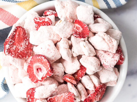 A bowl filled with cereal pieces and dried strawberries, reminiscent of a delightful strawberry puppy chow recipe, is dusted in white powder. It rests on a white marble surface, with a striped cloth in the background.