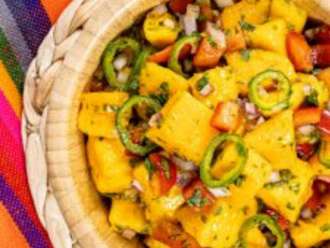 A woven basket brimming with a vibrant mango salsa recipe. The salad features diced mango, sliced green peppers, onions, and herbs. It rests on a colorful, striped cloth, crafting a festive and appetizing presentation.