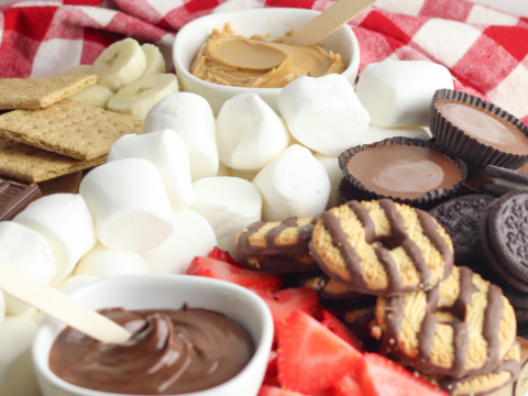 Experience the ultimate smores charcuterie board recipe idea with a wooden platter featuring marshmallows, strawberries, chocolate-covered cookies, peanut butter cups, dipped cookies, and graham crackers. Complete your indulgence with bowls of chocolate and peanut butter spread on a red and white checkered cloth backdrop.