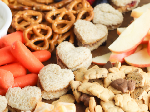 A delightful toddler charcuterie board featuring heart-shaped sandwiches, pretzels, cheese cubes, baby carrots, strawberries, blueberries, apple slices, and animal crackers.