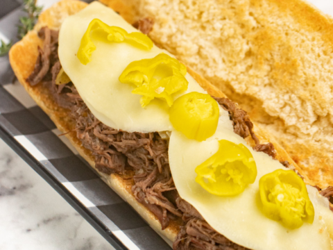 A sandwich on a checkered platter, reminiscent of an Italian Beef Hoagies Recipe, features shredded beef topped with two slices of cheese and banana pepper slices, served on a toasted sub roll. A sprig of herbs decorates the platter.