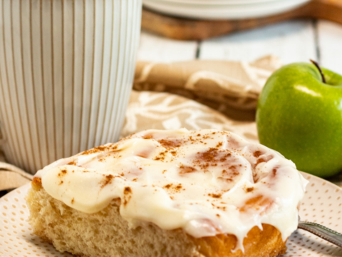 A close-up of an apple cinnamon roll with cream cheese frosting on a plate, dusted with cinnamon. Next to it is a green apple, a cup of coffee, and additional cinnamon sticks. Plates with more rolls are in the background on a wooden surface.