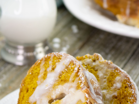 Slices of pumpkin Bundt cake drizzled with icing and dusted with powdered sugar are on a plate. A glass jug filled with milk is in the background, alongside the remaining pumpkin cream cheese coffee cake on a white serving dish.