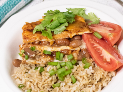 A white bowl of rice topped with sliced tomatoes, a golden-brown omelette, chopped green onions, and cilantro. The scene evokes a cozy fall vibe, perfect for enjoying while prepping Halloween treats like spooky Dunkaroo dip. A fork rests on the table beside a teal and white checkered cloth.
