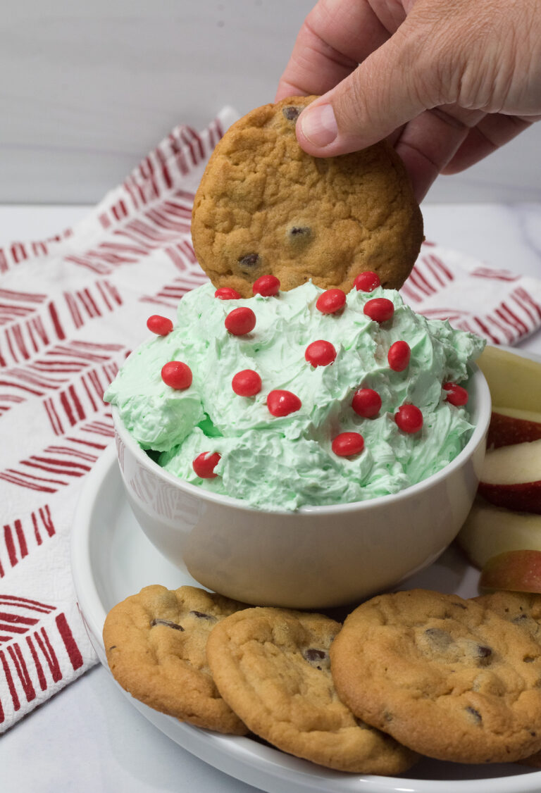 A hand dips a chocolate chip cookie into a bowl of Sweet Grinch Dip, a light green frosting topped with red candies. The bowl rests on a plate with more cookies and apple slices. A red and white patterned cloth is in the background, adding to the festive flair.