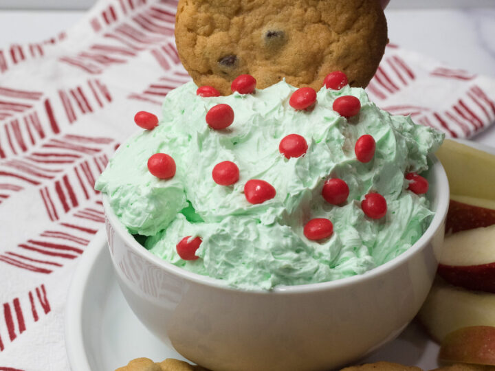 A hand dips a chocolate chip cookie into a bowl of Sweet Grinch Dip, a light green frosting topped with red candies. The bowl rests on a plate with more cookies and apple slices. A red and white patterned cloth is in the background, adding to the festive flair.